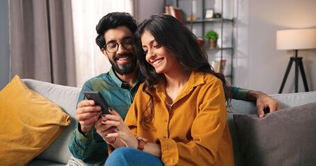 Portrait of cheerful positive young lovely couple smiling spending time together at home sitting on sofa typing on smartphone, searching internet using social network app on cellphone, family concept