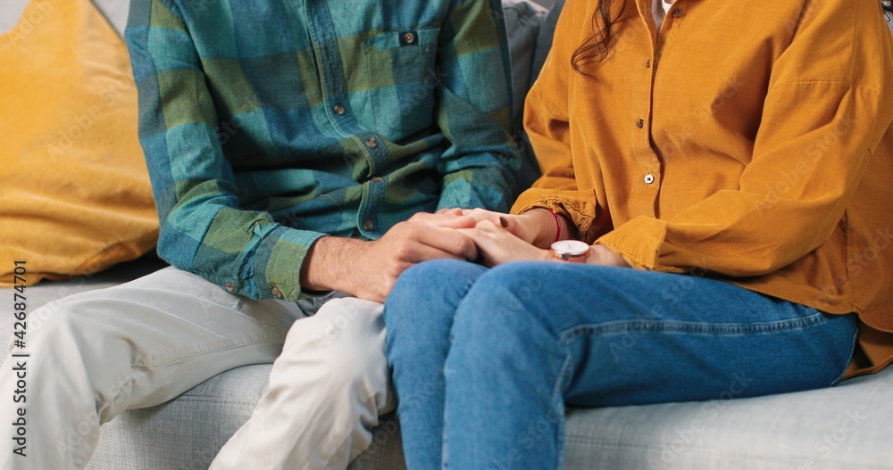 Close up of cheerful Hindu young couple man and woman in love in good mood speaking and laughing sitting on sofa in room at home, love and care, holding hands together, relationships concept Close up of cheerful Hindu young couple man and woman in love in good mood speaking and laughing sitting on sofa in room at home, love and care, holding hands together, relationships concept