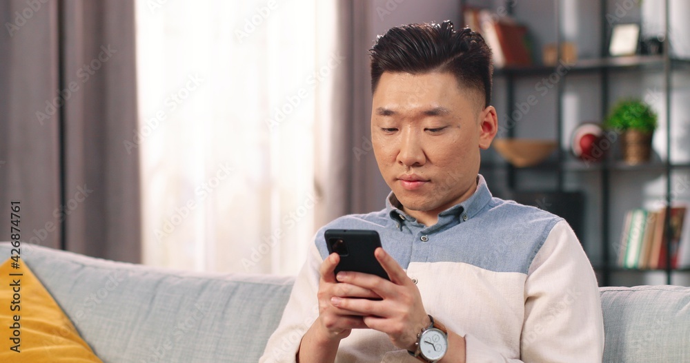 Young Asian joyful male resting at home sitting on couch in modern room texting online on smartphone. Man typing on mobile phone in good mood and smiling, social network, close up portrait concept Young Asian joyful male resting at home sitting on couch in modern room texting online on smartphone. Man typing on mobile phone in good mood and smiling, social network, close up portrait concept