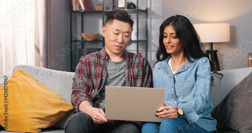 Portrait of cheerful positive mixed-race young couple wife and husband video chatting on laptop speaking on online call while sitting in cozy room on sofa, communication internet, home concept