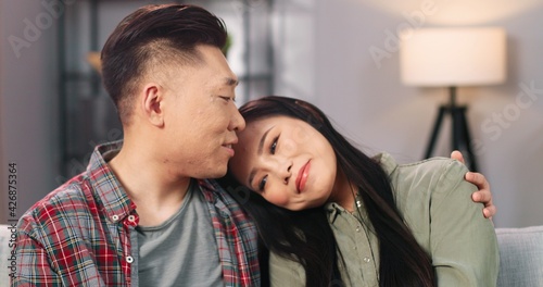 Close up of happy Asian young beautiful woman speaking with husband while sitting on couch in cozy room in apartment. Cheerful married couple in love spending time together in positive mood at home © VAKSMANV