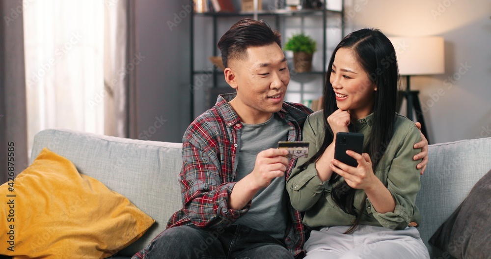 Portrait of Asian cheerful young couple male and female sitting on sofa in living room shopping online on smartphone, buying on sales using credit card, hugging and smiling, buyer concept Portrait of Asian cheerful young couple male and female sitting on sofa in living room shopping online on smartphone, buying on sales using credit card, hugging and smiling, buyer concept