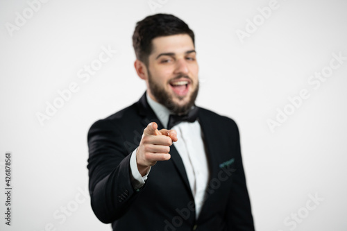 Charming man in tuxedo points index finger into camera lens and winks, flirts. Hey, you. gentleman with beard in holiday festive suit on white background.