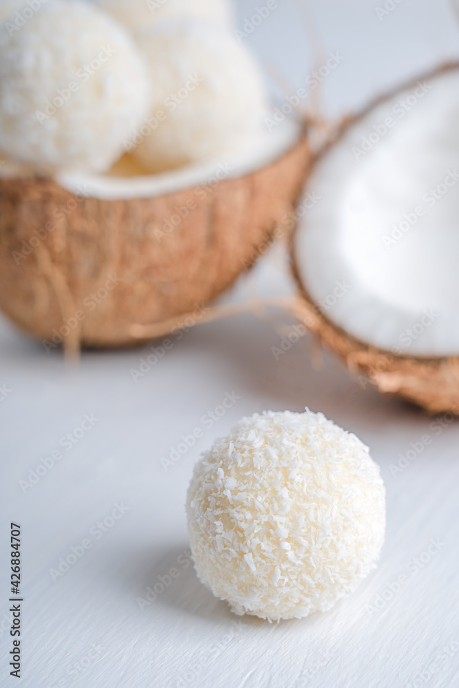 Coconut vegan truffles or homemade vegetarian energy balls with a filling of condensed milk and curd cheese served in halved fresh coconut on white wooden table ready for eating. Vertical image