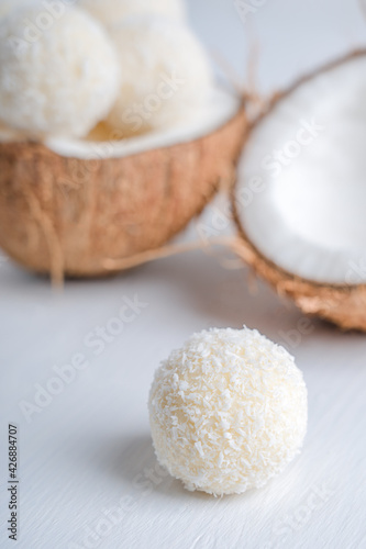 Coconut vegan truffles or homemade vegetarian energy balls with a filling of condensed milk and curd cheese served in halved fresh coconut on white wooden table ready for eating. Vertical image