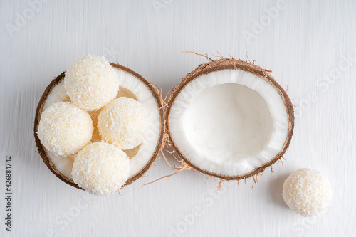 Top view of coconut truffles or homemade vegetarian energy balls with a filling of sweetened shredded coconut and curd cheese served in halved fresh coconut on white wooden table. Horizontal image