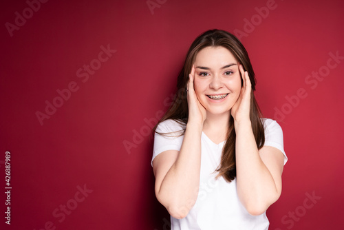 A beautiful girl in a white T-shirt with long hair puts her hands to her face and smiles broadly in braces © DmitryStock