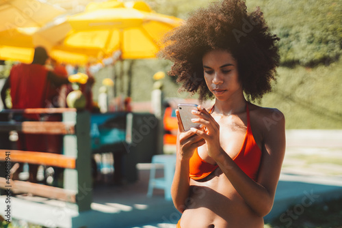 Portrait of a young ravishing African-American female with fluffy afro hair and in a swimsuit sending a message to her new boyfriend via smartphone while standing near the beach kiosk on a summer day photo