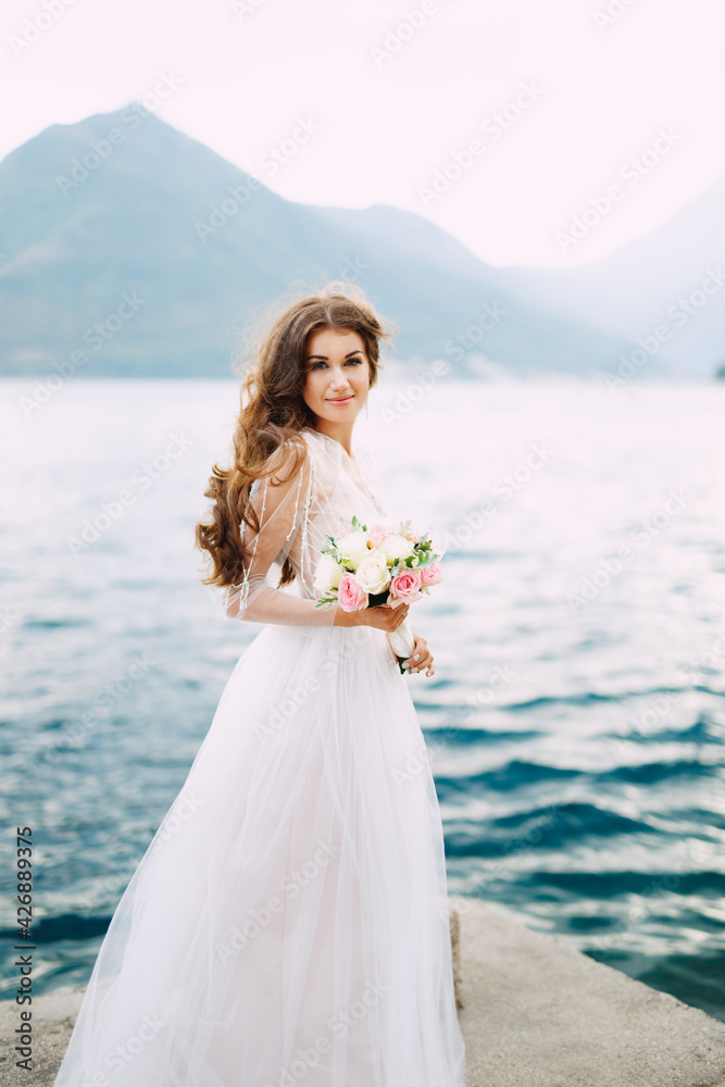 The bride holds a bouquet of roses in her hands and stands on the pier in the Bay of Kotor