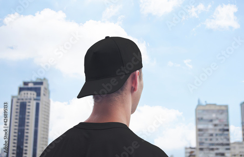 Young man in a white baseball cap and white t-shirt on background of the city and blue sky with clouds, baseball cap mock up