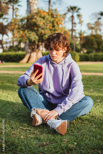 Angry teenager wearing a purple hoodie looking at a mobile phone in the park. She is sitting in the grass. © Pablo Rasero