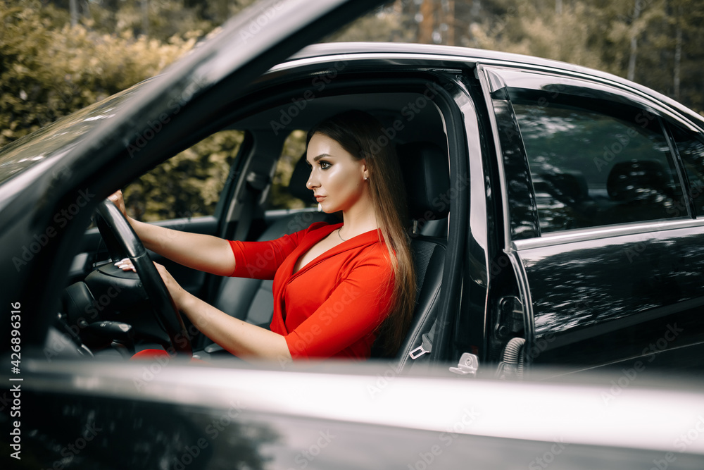 A beautiful young girl in a red overalls sits behind the wheel of a black car on an empty road in the forest