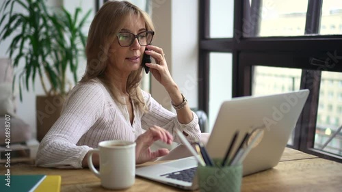 Businesswoman talking on phone and sitting at table with laptop in home office.