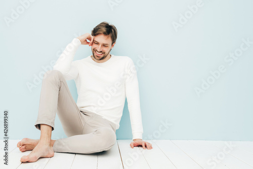 Portrait of handsome smiling hipster lumbersexual businessman model wearing casual white sweater and trousers. Fashion stylish man posing against light blue wall in studio