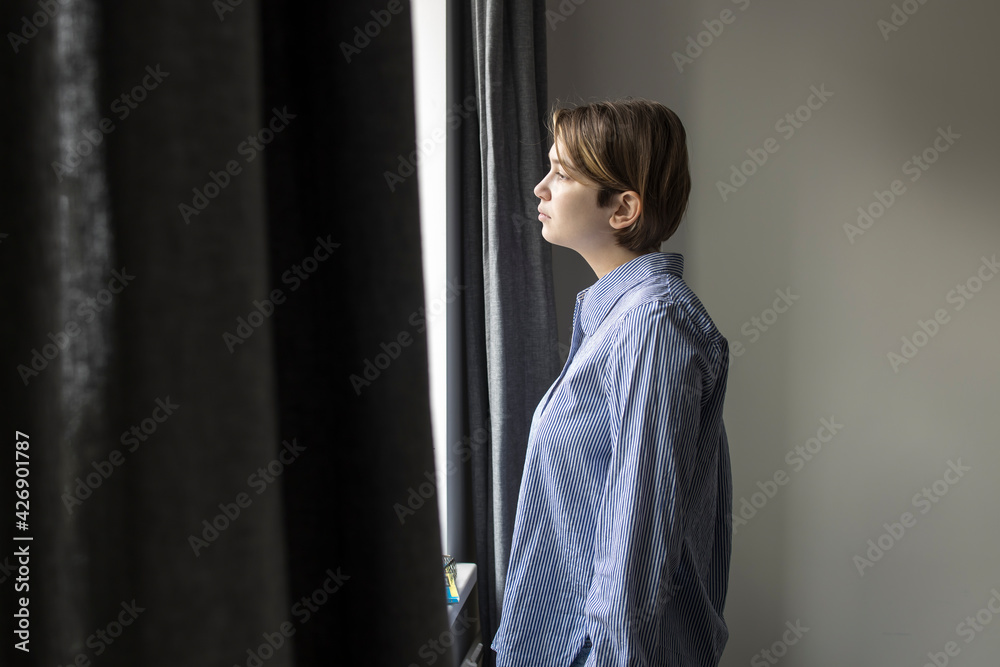 Fototapeta premium A pensive girl with a short hair nineteen years old in a white shirt with blue stripes looks out the window