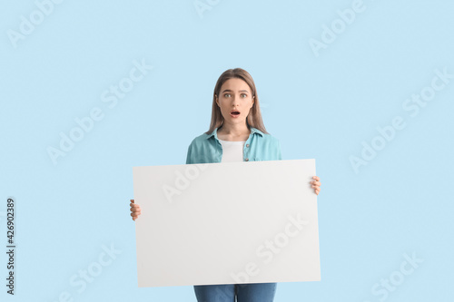 Surprised young woman with blank poster on color background