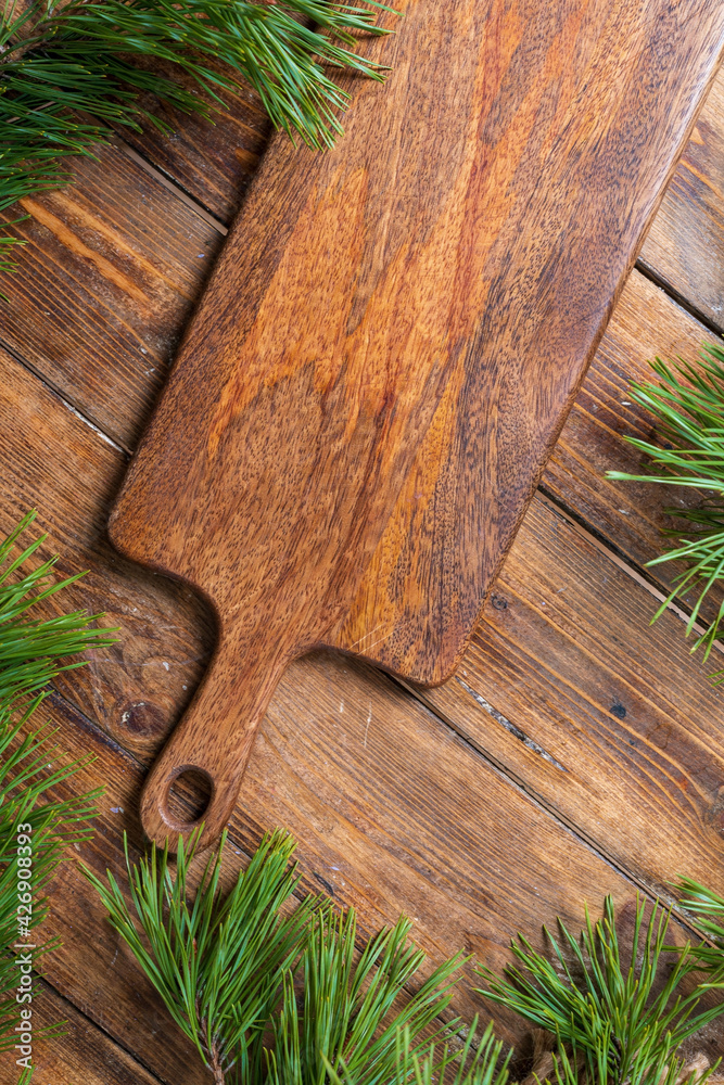 Cutting board on a wooden table background surrounded by pine branches, top view. Vertical. Cutting board on a wooden table background surrounded by pine branches, top view. Vertical.