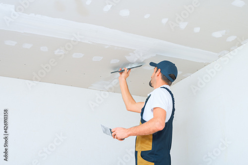 A uniformed worker applies putty to the drywall ceiling. Putty of joints of drywall sheets. photo