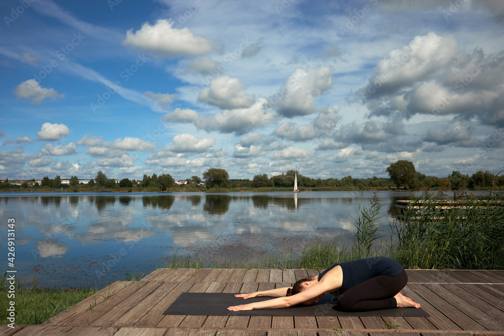 Fototapeta premium Slim young woman practicing yoga on the wooden pier near lake.