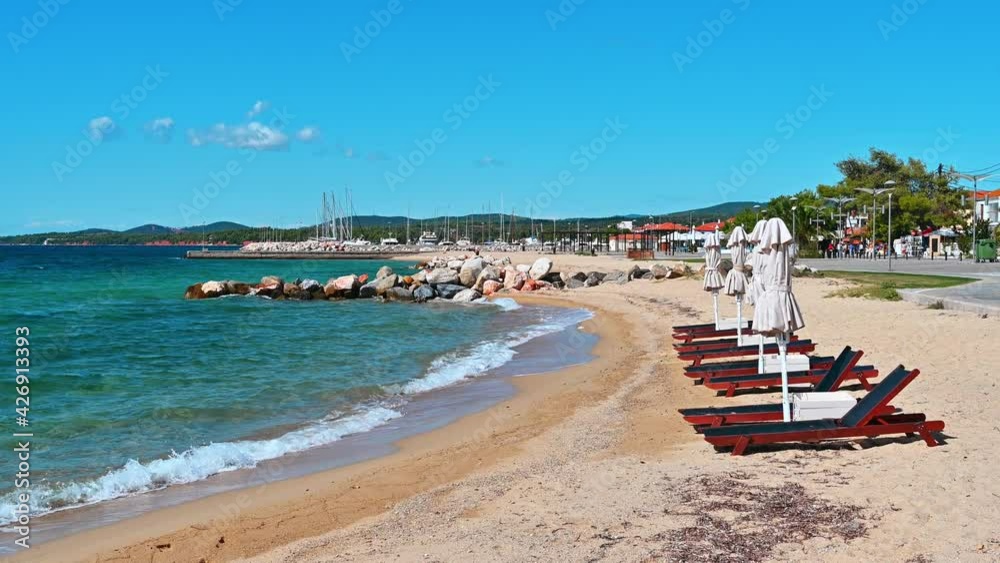 Beach on Aegean sea coast with closed umbrellas and sunbeds, rocks in the water, embankment street, sea port and green hills in the distance in Nikiti, Greece Beach on Aegean sea coast with closed umbrellas and sunbeds, rocks in the water, embankment street, sea port and green hills in the distance in Nikiti, Greece