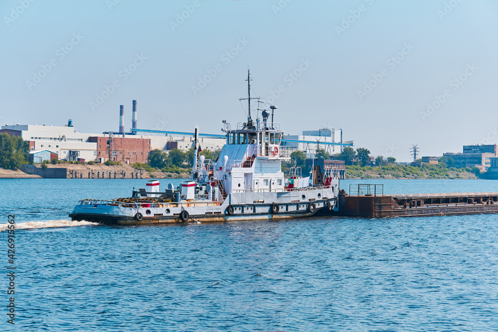 towboat pushes dry bulk cargo barge on the river past the industrial landscape