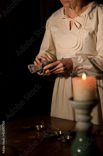 Eine Frau steht im alten Haus und putzt mit dem Lappen beim Kerzenlicht das Geschirr ab. Sie ist angezogen wie eine Bäuerin mit dem Kopftuch, einem alten Kleid und einer Schürze. 