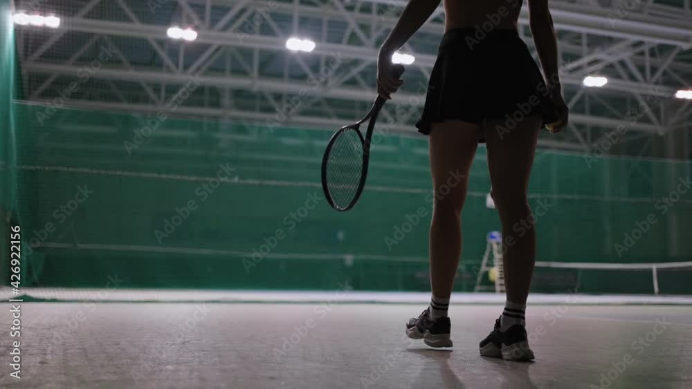 sporty woman is walking on tennis court indoors, throwing ball and catching, rear view, professional sport workout