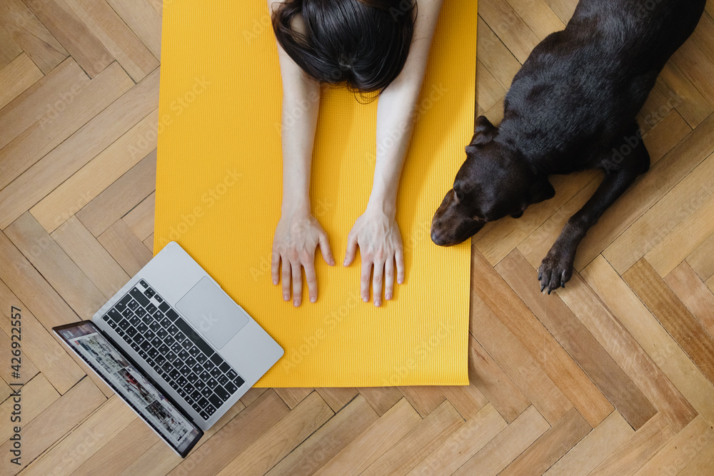 European caucasian woman does yoga and meditation in her home, home calming yoga on a yellow mat. woman prepares for yoga class online with a laptop, talking mat and doing stretching, millennial woman - obrazy, fototapety, plakaty 