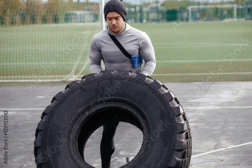 Strong white man lifts a large wheel on an outdoor sports field. The concept of a strong minded athlete going to his goal