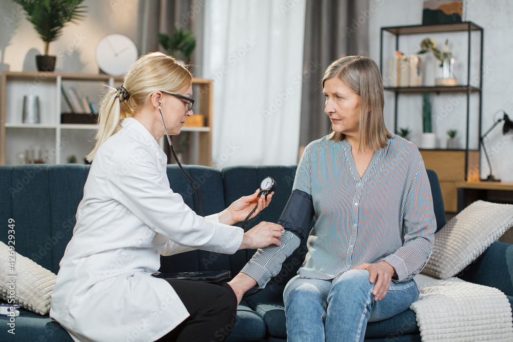 Obraz premium Seniors hypertension concept. Doctor measuring blood pressure to patient at home. Female professional doctor examining elderly old woman patient, measuring arterial blood pressure using tonometer.