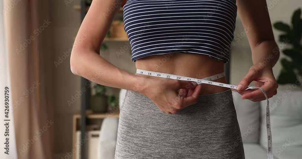Close up sporty young fit woman with tightened abdominal muscles in activewear measuring waist line with tape after fat burn workout at home, healthcare slimming weight loss, dietary lifestyle concept