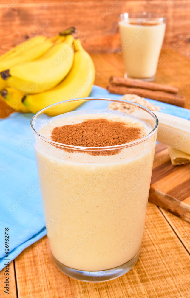 banana and oat smoothie with cinnamon powder on top, on one side cinnamon sticks and a peeled banana on a wooden table and a background of white wooden planks. banana and oat smoothie with cinnamon powder on top, on one side cinnamon sticks and a peeled banana on a wooden table and a background of white wooden planks.