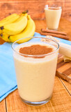 banana and oat smoothie with cinnamon powder on top, on one side cinnamon sticks and a peeled banana on a wooden table and a background of white wooden planks.