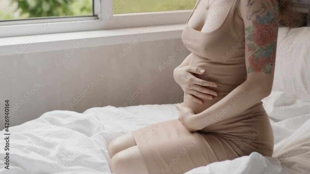 Happy pregnant woman stroking belly near window indoors. Pretty expectant mother sitting in light room. Pregnant woman smiling to unborn baby. Future mom petting her stomach. Happy pregnant woman stroking belly near window indoors. Pretty expectant mother sitting in light room. Pregnant woman smiling to unborn baby. Future mom petting her stomach.