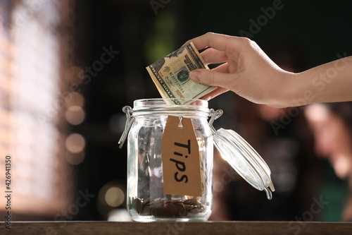 Woman putting tips into glass jar on wooden table indoors, closeup photo