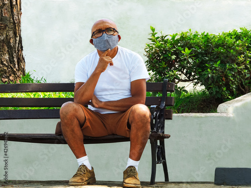 Elderly man wearing face mask for protection against coronavirus sitting on a bench next to a garden and a white wall background . Thoughtful bald old man sitting alone during covid-19 pandemic