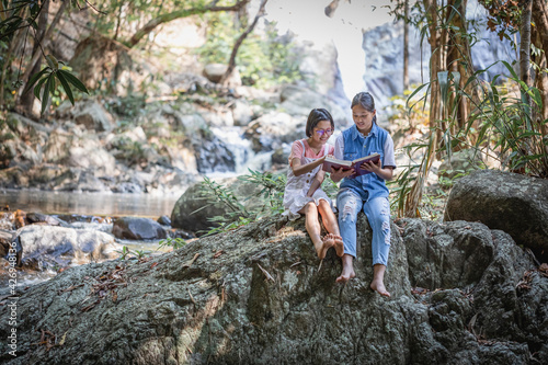 Cute two children girls are reading a book sitting on stone in forest .