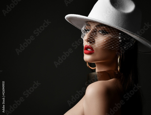 Portrait of rich gorgeous woman in wide-brimmed white hat with veiling and massive gold collar and earrings. stands with bared shoulders over black background. Stylish look and sexual games concept