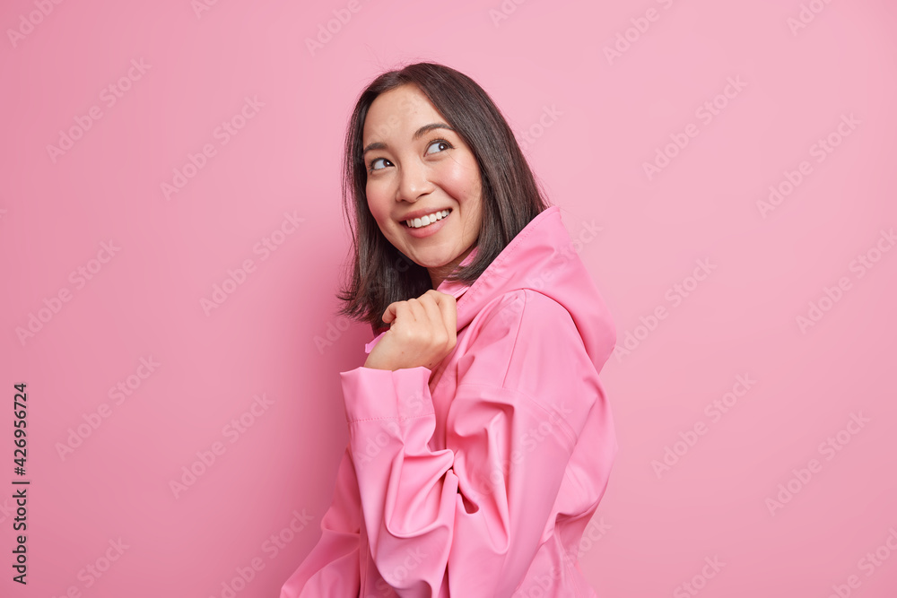Pretty glad brunette Asian woman stands half turned against pink studio background has good mood wears stylish jacket with hood thinks about something pleasant poses happy indoor. Emotions concept Pretty glad brunette Asian woman stands half turned against pink studio background has good mood wears stylish jacket with hood thinks about something pleasant poses happy indoor. Emotions concept