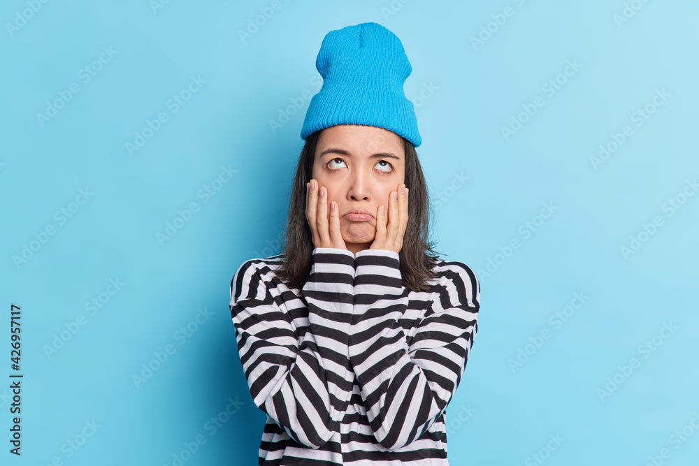 Half length shot of bored stressed unamused teenage girl keeps palms on cheeks concentrated above with displeased reluctant expression poses against blue background dressed in casual clothes Half length shot of bored stressed unamused teenage girl keeps palms on cheeks concentrated above with displeased reluctant expression poses against blue background dressed in casual clothes