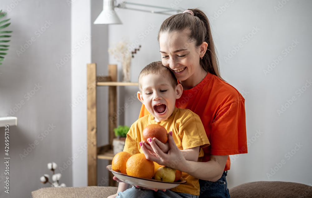 Cheerful mom and child are fooling around, laughing with a plate of fruits in their hands. Concept of a happy and healthy family, pastime with children and a bright mood Cheerful mom and child are fooling around, laughing with a plate of fruits in their hands. Concept of a happy and healthy family, pastime with children and a bright mood