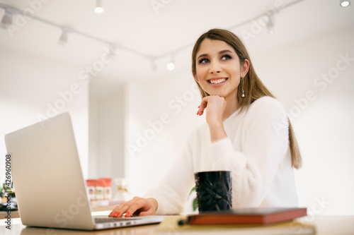 Beautiful Caucasian woman thinking about something while sitting with notebook in a minimal cafe bar. Charming female freelancer thinking about new ideas during work on laptop drinking coffee © arthurhidden