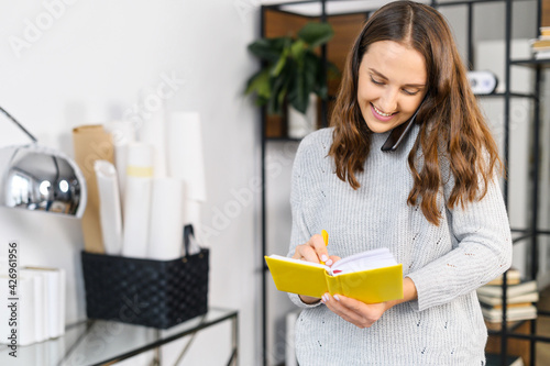 Cheerful woman has phone conversation and writes down into notebook, standing in the office, multitasking female office employee