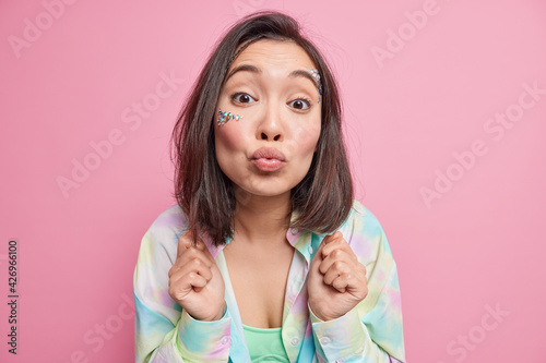 Lovely romantic brunette Asian woman looks tenderly at camera keeps lips folded in mwah wants to kiss you wears colorful shirt poses against pink background bends forward makes kissing face.