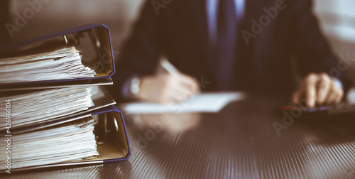 Binders with papers are waiting to be processed by man accountant in blue blazer staying at home during covid pandemic. Taxes and audit concept. Internal Revenue Service inspector checking financial