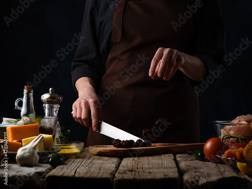 Chef cuts blackberries for cooking meat or salad on the background of ingredients.Various cheeses, garlic, tomatoes and spices.Culinary recipes