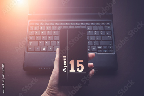 April 15th. Day 15 of month, Calendar date. Hand Holding Mobile Phone on Laptop Computer dark background with sunshine. Spring month, day of the year concept.