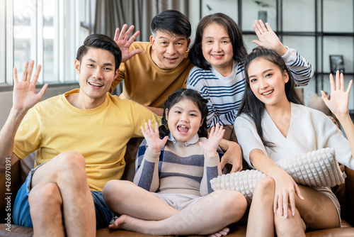 family portrait asian multi gneration  parent grandparent and grandchild sit relax together smile hand wave to say hi look at camera on sofa at living room home interior background