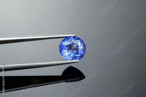 Natural oval portuguese faceted rich purplish blue color crystal clean, flawless tanzanite semoprecious gemstone holded in tweezers. Gray reflective background.