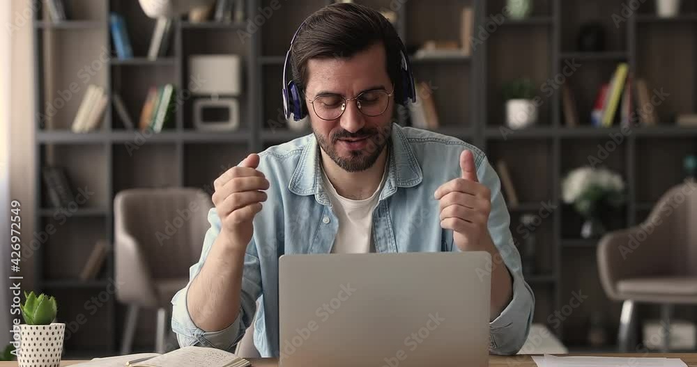 Happy young man in eyeglasses wearing wireless headphones, enjoying online video call using computer zoom application. Smiling 30s manager communicating distantly with colleagues at web camera meeting